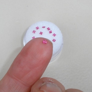 May include: A collection of small, square-cut, vibrant pink gemstones. One individual pink gemstone is held up, while several others are arranged on a circular white foam insert inside a small, clear, round plastic storage container.