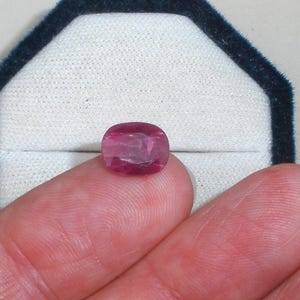 May include: A cushion-cut gemstone with a deep magenta-pink hue and a faceted surface that reflects light. The stone is held against a white velvet-lined display box with a dark blue border, showcasing its translucent clarity and polished finish.