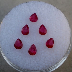 May include: Six pear-shaped ruby gemstones on a white background. The gemstones are arranged in a triangular pattern.