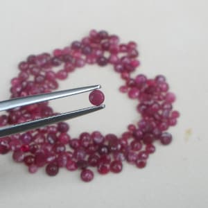 May include: A single, round, deep red gemstone held by silver tweezers, with a scattering of similar gemstones in the background. The gemstones are a rich, dark red color, and the tweezers are silver.