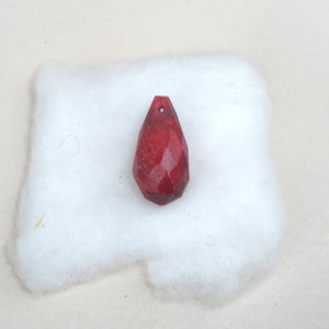 May include: A deep red, faceted teardrop-shaped pendant with a small hole at the top for suspension. The surface of the pendant features multiple angular cuts that create a geometric, multi-faceted appearance, reflecting light across its dark crimson surface.