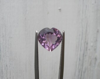 Könnte beinhalten: Ein herzförmiger, facettierter Amethyst-Edelstein, der von einer Metallpinzette gehalten wird. Der Edelstein hat eine hellviolette Farbe mit dunkleren violetten Facetten. Die Pinzette ist silberfarben und der Hintergrund ist hellgrau.