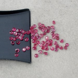 Może przedstawiać: Kolekcja różowych kamieni szlachetnych, prawdopodobnie rubinów, rozrzuconych na czarnej tacy i jasnoszarym podłożu.