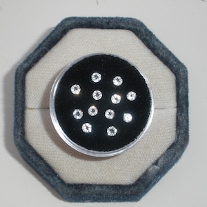 Può includere: Una collezione di piccole gemme rotonde e trasparenti esposte su una superficie di velluto nero all'interno di un contenitore di plastica trasparente. Il contenitore è posto all'interno di un portagioie ottagonale blu scuro.
