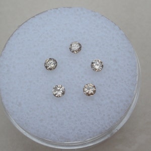 Op de afbeelding: Vijf kleine, ronde, heldere diamanten met een lichte bruine tint worden getoond op een wit, getextureerd oppervlak.