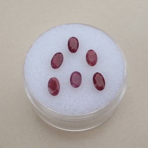 May include: Six oval-shaped, red gemstones in a white, round container.