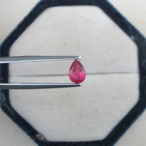 May include: A pear-shaped, deep red ruby gemstone held by tweezers against a white background.