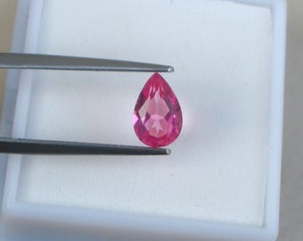 May include: A faceted, pear-shaped, pink gemstone held by tweezers. The gem is a vibrant shade of pink, with multiple facets catching the light. It is set against a white background within a small, open, white plastic container.