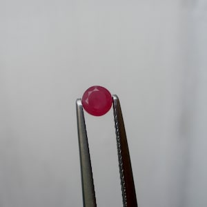Pode incluir: Um rubi vermelho redondo e facetado é segurado por uma pinça prateada contra um fundo branco. O rubi é de uma cor vermelha profunda e saturada e parece polido.