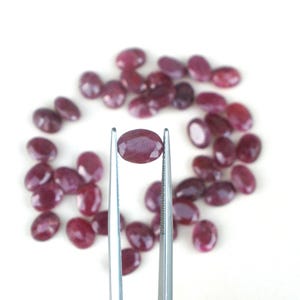 May include: A collection of oval-shaped, deep red gemstones. One gemstone is held by silver tweezers, showcasing its polished surface. The gems are scattered on a white background, highlighting their color and shape. Ideal for jewelry making.