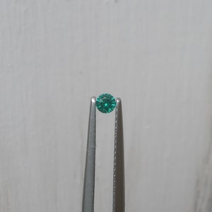 May include: A bright green gemstone held in tweezers against a white background.