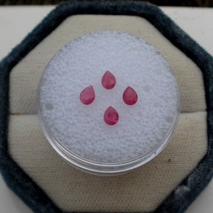 Op de afbeelding: Vier roze peervormige edelstenen worden getoond in een container met een witte schuimvoering. De container is rond en heeft een doorzichtig plastic deksel.