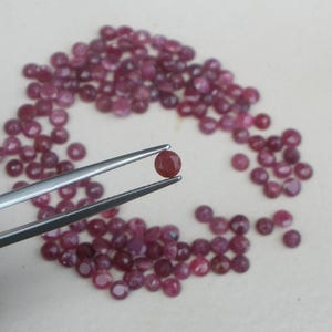 Puede incluir: Colección de pequeñas gemas de rubí redondas y facetadas. Una gema es sostenida por unas pinzas plateadas, con muchas más dispersas sobre una superficie clara. Los rubíes son de un color rojo intenso.