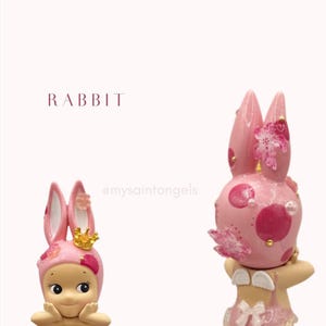 May include: Two pink rabbit figurines with floral accents and gold crowns. One figurine has a smiling face and is wearing a pink bunny hat. The other figurine has a pink polka-dotted head and is wearing a pink swimsuit with angel wings.