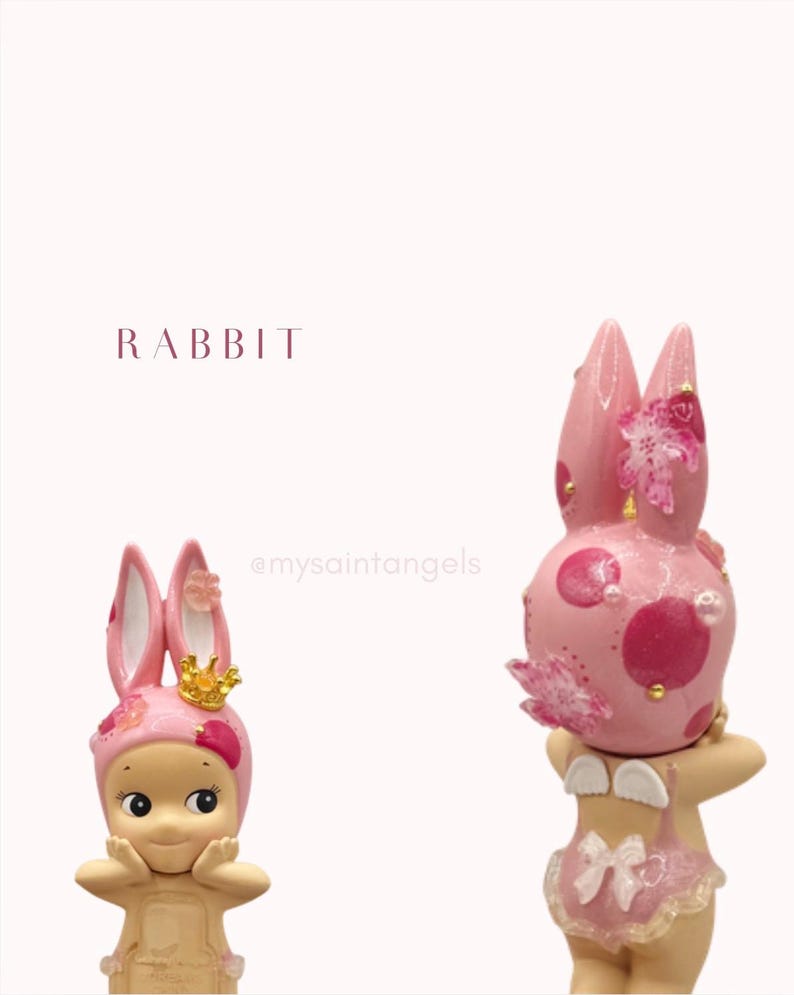 May include: Two pink rabbit figurines with floral accents and gold crowns. One figurine has a smiling face and is wearing a pink bunny hat. The other figurine has a pink polka-dotted head and is wearing a pink swimsuit with angel wings.