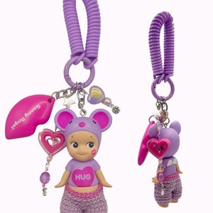 Puede incluir: Un llavero morado con una figura de rat&oacute;n que lleva una camiseta con la palabra "HUG". El llavero incluye un dije de labios rosa, un dije de coraz&oacute;n y un cord&oacute;n en espiral. La palabra "Mouse" est&aacute; escrita en la esquina superior derecha.
