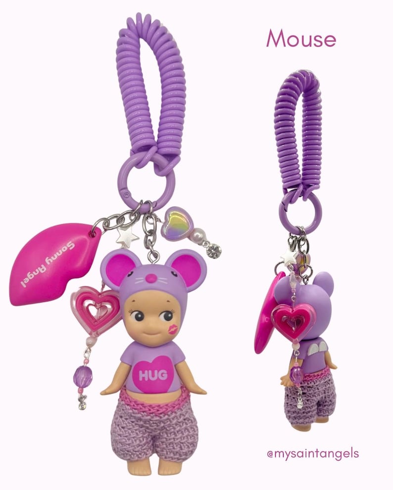 Puede incluir: Un llavero morado con una figura de rat&oacute;n que lleva una camiseta con la palabra "HUG". El llavero incluye un dije de labios rosa, un dije de coraz&oacute;n y un cord&oacute;n en espiral. La palabra "Mouse" est&aacute; escrita en la esquina superior derecha.