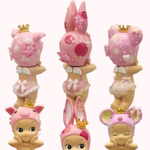May include: A collection of six pink figurines, each adorned with a crown and animal-themed headwear. The figurines include pig, bunny, and mouse designs, with delicate floral and polka dot patterns. The text "@mysaintangels" is in the top left corner.