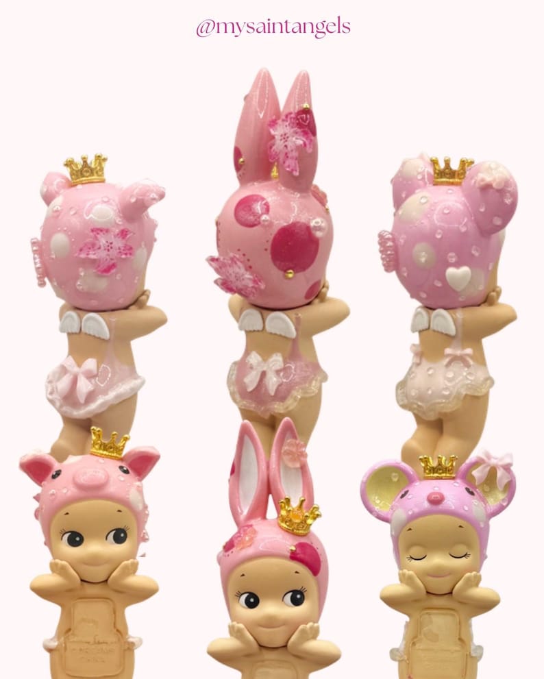 May include: A collection of six pink figurines, each adorned with a crown and animal-themed headwear. The figurines include pig, bunny, and mouse designs, with delicate floral and polka dot patterns. The text "@mysaintangels" is in the top left corner.