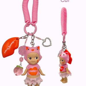 Puede incluir: Llavero rosa con una mu&ntilde;eca con tem&aacute;tica de gato, con una correa de mu&ntilde;eca en espiral rosa. La mu&ntilde;eca lleva una camiseta rosa con "SWEET", una falda rosa y una capucha con orejas de gato. Incluye un dije con el texto "Sonny Angel".