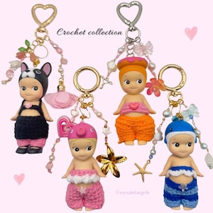 Könnte beinhalten: Vier Schlüsselanhänger mit kleinen Puppenfiguren mit Häkeloutfits und Accessoires. Die Puppen haben verschiedene Hüte und Outfits in Schwarz, Rosa, Orange und Blau. Jeder Schlüsselanhänger hat einen herzförmigen Verschluss und dekorative Perlen. Der Text lautet "Crochet collection".