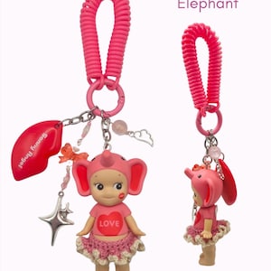 Puede incluir: Un llavero rosa con tem&aacute;tica de elefante con una peque&ntilde;a figura que lleva un sombrero y una falda de elefante rosa. La figura tiene un coraz&oacute;n con la palabra "LOVE" en su camisa. El llavero incluye un dije de labio rojo y un dije de estrella.