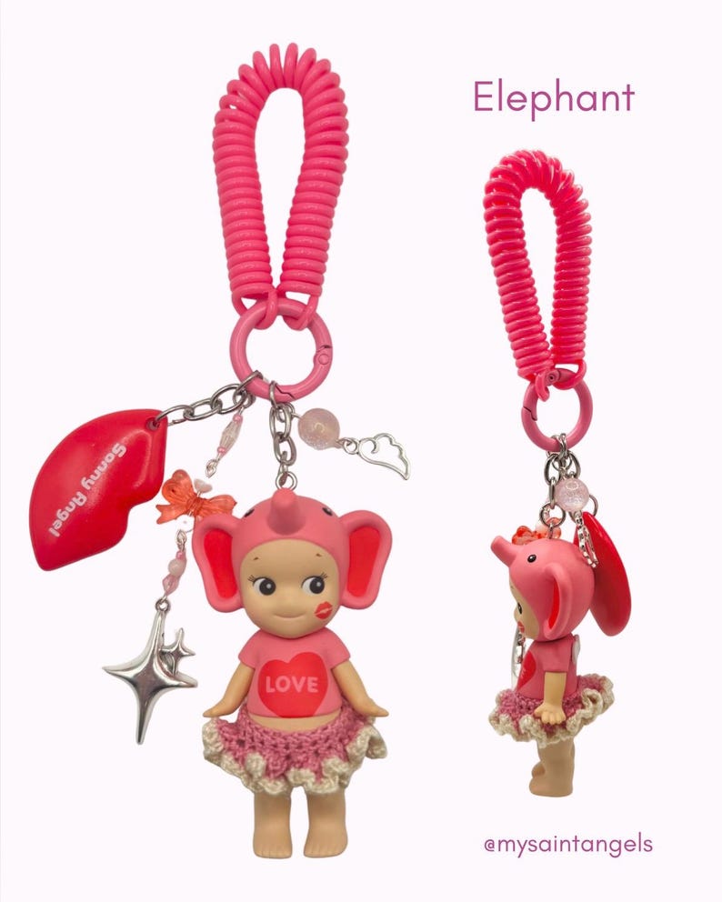 Puede incluir: Un llavero rosa con tem&aacute;tica de elefante con una peque&ntilde;a figura que lleva un sombrero y una falda de elefante rosa. La figura tiene un coraz&oacute;n con la palabra "LOVE" en su camisa. El llavero incluye un dije de labio rojo y un dije de estrella.