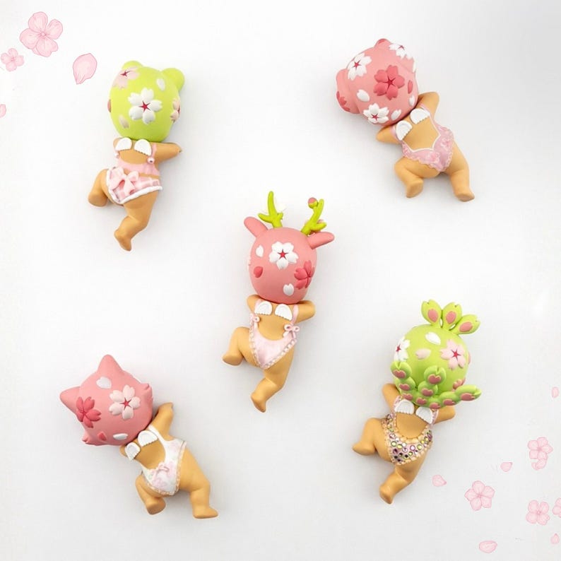 May include: Five baby-shaped figurines with floral headpieces and swimsuits. The figurines are in pink and green, with white accents. The background is white. The figurines are decorated with flowers.