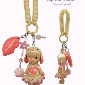 Puede incluir: Un llavero Lop Ear Rabbit con una mu&ntilde;eca gui&ntilde;ando un ojo que lleva una camiseta blanca con un coraz&oacute;n rojo que dice "CUTIE". La mu&ntilde;eca tiene un tut&uacute; rosa y orejas de conejo. El llavero incluye un dije de labio rosa, un dije de pata y cuentas rosas. El texto en la imagen dice "Secret Sonny".