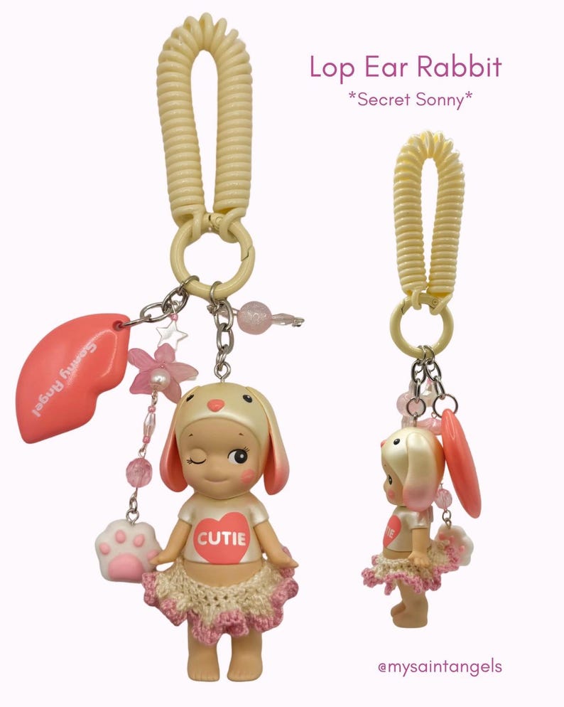 Puede incluir: Un llavero Lop Ear Rabbit con una mu&ntilde;eca gui&ntilde;ando un ojo que lleva una camiseta blanca con un coraz&oacute;n rojo que dice "CUTIE". La mu&ntilde;eca tiene un tut&uacute; rosa y orejas de conejo. El llavero incluye un dije de labio rosa, un dije de pata y cuentas rosas. El texto en la imagen dice "Secret Sonny".