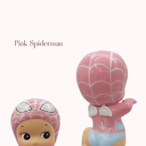 May include: Two pink figurines of a baby dressed as Spiderman. The figurines have a pink head with white web detailing, a light blue diaper, and small white wings. The text "Pink Spiderman" is in the image.