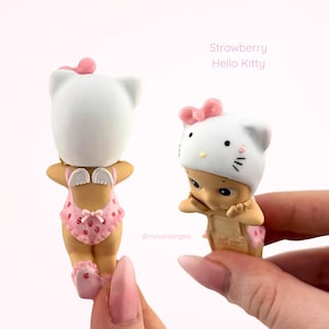 May include: Two small figurines featuring Hello Kitty wearing a white hood with a pink bow. One figurine shows the back with white angel wings and a pink strawberry-patterned leotard, while the other shows the front. Text reads Strawberry Hello Kitty.