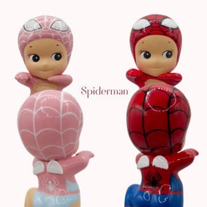 May include: Two figurines of a baby dressed as Spiderman. One is pink with a light blue diaper, the other is red and blue. The text "Spiderman" is in the centre of the image.