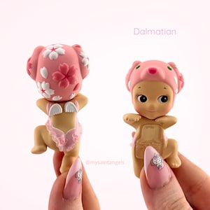 May include: Two small figurines, one viewed from the back, the other from the front. Both are wearing pink head coverings and outfits. The text "Dalmatian" is above the figurines. The figurines are held by a person with a pink manicure.
