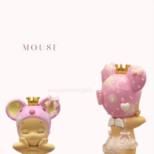 May include: Two pink mouse figurines with golden crowns. The figurines have a light pink and white polka dot design on their heads and ears. The word "MOUSE" is printed in the upper left corner.