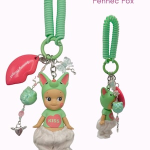 Puede incluir: Dos llaveros Sonny Angel Fennec Fox. Cada llavero presenta una peque&ntilde;a figura con un gorro de zorro verde y una camiseta con un coraz&oacute;n que dice "Kiss". Los llaveros tienen una espiral verde, un dije de labio rosa y otros dijes decorativos.