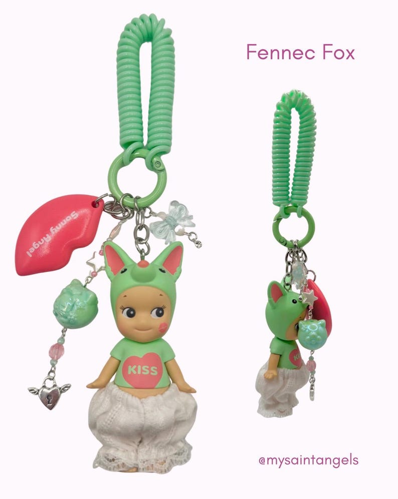Puede incluir: Dos llaveros Sonny Angel Fennec Fox. Cada llavero presenta una peque&ntilde;a figura con un gorro de zorro verde y una camiseta con un coraz&oacute;n que dice "Kiss". Los llaveros tienen una espiral verde, un dije de labio rosa y otros dijes decorativos.