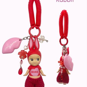 Puede incluir: Un charm rojo con tem&aacute;tica de conejo con una mu&ntilde;eca vestida con un atuendo rojo y orejas de conejo. La mu&ntilde;eca tiene un coraz&oacute;n con "XOXO" en su camisa. Un charm en forma de labio rosa y un llavero de espiral rojo completan el dise&ntilde;o.