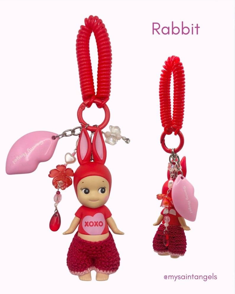 Puede incluir: Un charm rojo con tem&aacute;tica de conejo con una mu&ntilde;eca vestida con un atuendo rojo y orejas de conejo. La mu&ntilde;eca tiene un coraz&oacute;n con "XOXO" en su camisa. Un charm en forma de labio rosa y un llavero de espiral rojo completan el dise&ntilde;o.