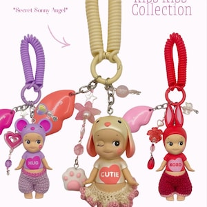 Puede incluir: Tres figuras de Sonny Angel de la colecci&oacute;n Kiss Kiss, cada una con un sombrero y atuendo tem&aacute;tico de animales. Las figuras est&aacute;n unidas a llaveros con dijes y cuentas decorativas. El texto "Kiss Kiss Collection" es visible.