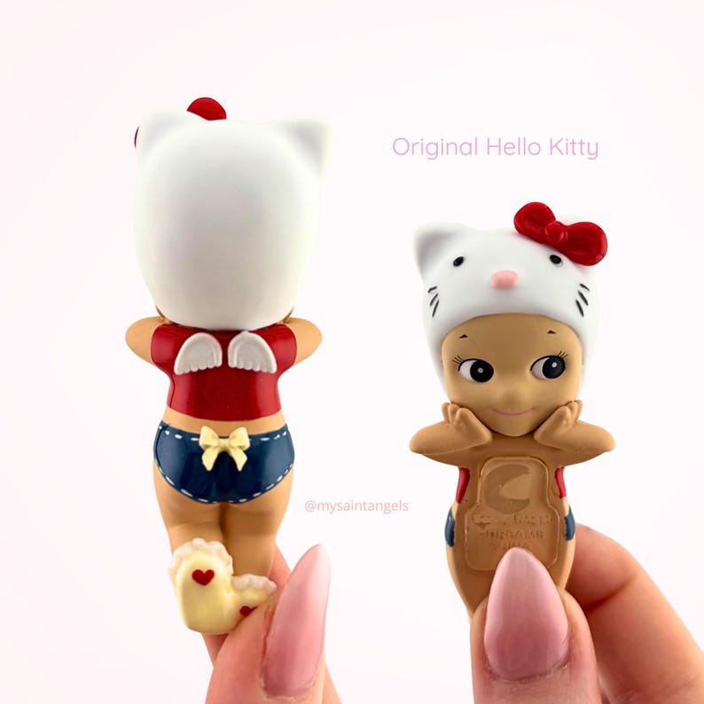 May include: Two Hello Kitty themed figurines featuring a tan-skinned character wearing a white cat hood with a red bow. One figure shows the back with white angel wings and blue shorts with a yellow bow, while the other shows the front with hands on cheeks.