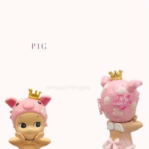 May include: Two figurines of cherubic figures, one with a pig hat and the other with a pig head, both wearing golden crowns. The figurines are pink and beige, with delicate details. The word "PIG" is written in a burgundy color.
