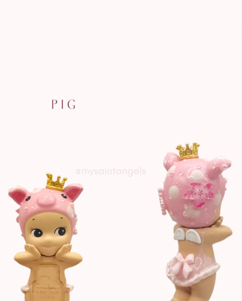May include: Two figurines of cherubic figures, one with a pig hat and the other with a pig head, both wearing golden crowns. The figurines are pink and beige, with delicate details. The word "PIG" is written in a burgundy color.