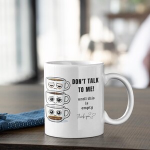 May include: White ceramic coffee mug with the text "DON'T TALK TO ME! until this is empty Thank you" and three stacked coffee cup illustrations with different facial expressions. The mug has a curved handle and is on a wooden surface.