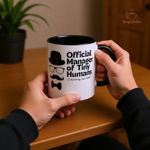 Può includere: Una tazza di ceramica bianca con interno e manico neri. La tazza presenta la scritta "Official Manager of Tiny Humans" con illustrazioni di cappello, occhiali, baffi e papillon. La tazza è tenuta da due mani.
