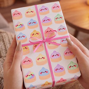 May include: A rectangular gift box wrapped in white paper featuring a repeating pattern of colourful, smiling cartoon dumplings in bamboo steamers, tied with a bright pink ribbon that is knotted into a bow at the centre of the package.