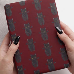 May include: A rectangular item wrapped in deep red paper featuring a repeating pattern of small black cats with yellow eyes and small black wings. The cats are depicted in a seated position, evenly spaced across the dark red background surface.