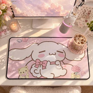 May include: A pink and white desk mat featuring a cartoon character with large ears, a bow, and a sad expression. The mat has a black border and includes a teddy bear, hearts, and a rose graphic. A computer monitor and accessories are in the background.