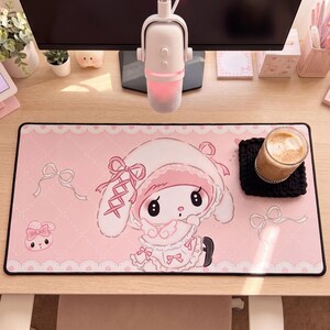 Pu&ograve; includere: Un tappetino da scrivania rettangolare con sfondo rosa chiaro, motivo trapuntato bianco, bordi in pizzo e illustrazioni di un coniglio bianco con cuffietta e vestito rosa, arricchito da piccoli fiocchi rosa e un secondo muso di coniglio.