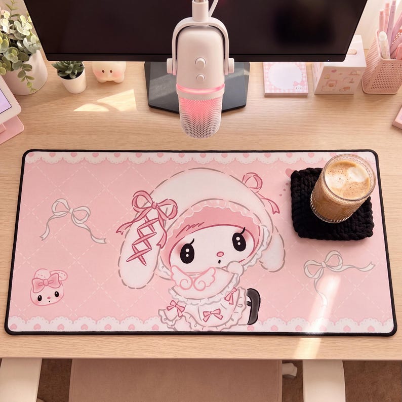 Pu&ograve; includere: Un tappetino da scrivania rettangolare con sfondo rosa chiaro, motivo trapuntato bianco, bordi in pizzo e illustrazioni di un coniglio bianco con cuffietta e vestito rosa, arricchito da piccoli fiocchi rosa e un secondo muso di coniglio.